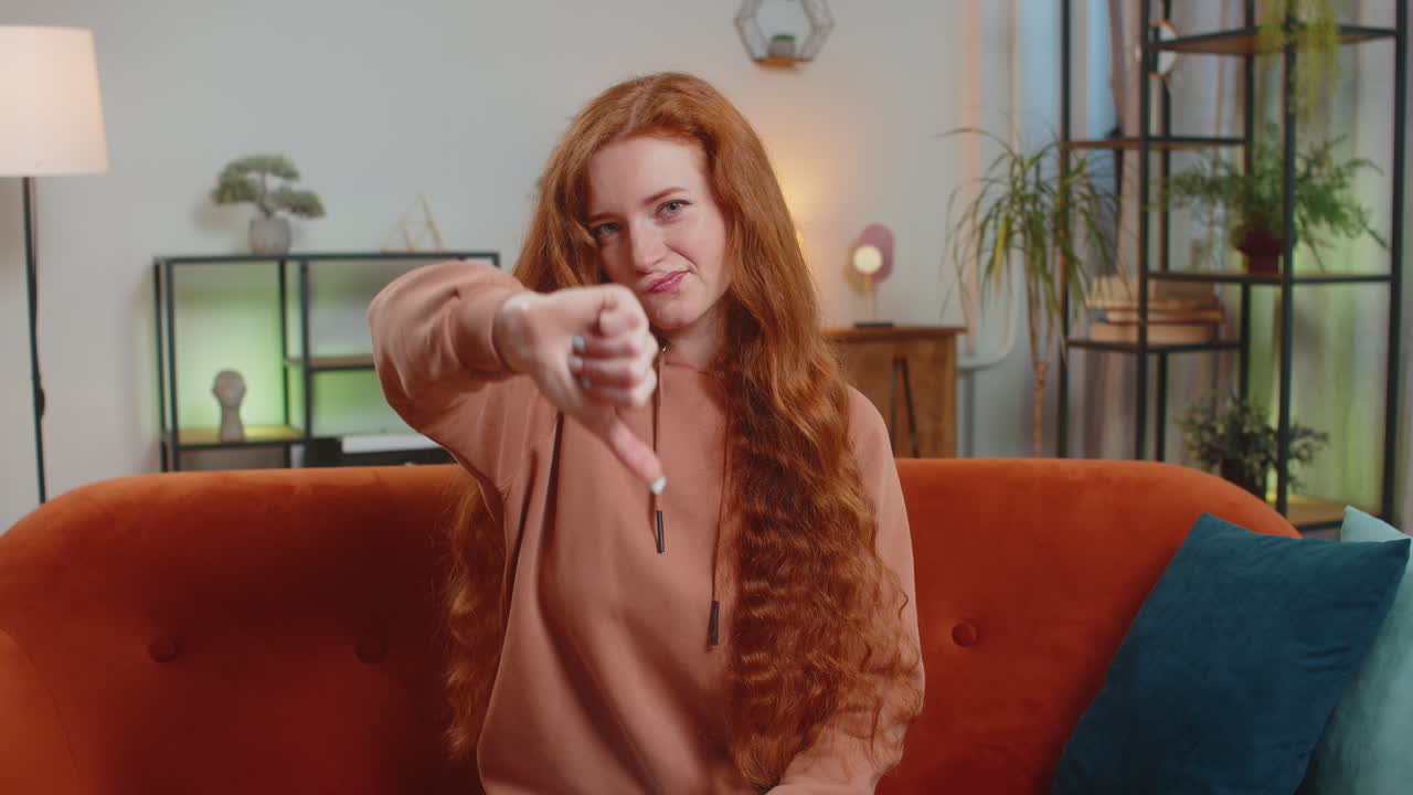 Upset woman showing thumb down sign gesture expressing discontent sitting on sofa couch at home