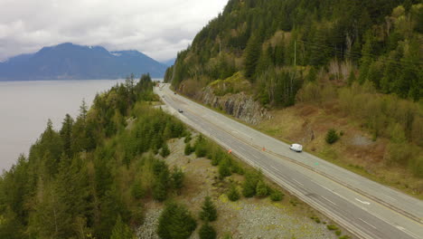 Vista-Aérea-De-Los-Automóviles-Que-Conducen-Por-Una-Carretera-Escénica-A-Lo-Largo-De-La-Hermosa-Costa-Del-Pacífico-En-La-Columbia-Británica