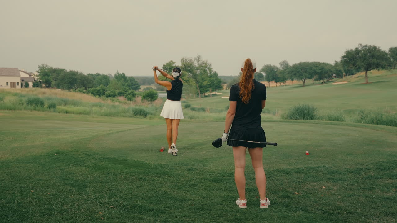 Una golfista colpisce con il driver dal tee al rallentatore, mentre la sua compagna di gioco osserva da dietro. Cattura la concentrazione, l'atletismo e il cameratismo su un campo da golf all'aperto e soleggiato.