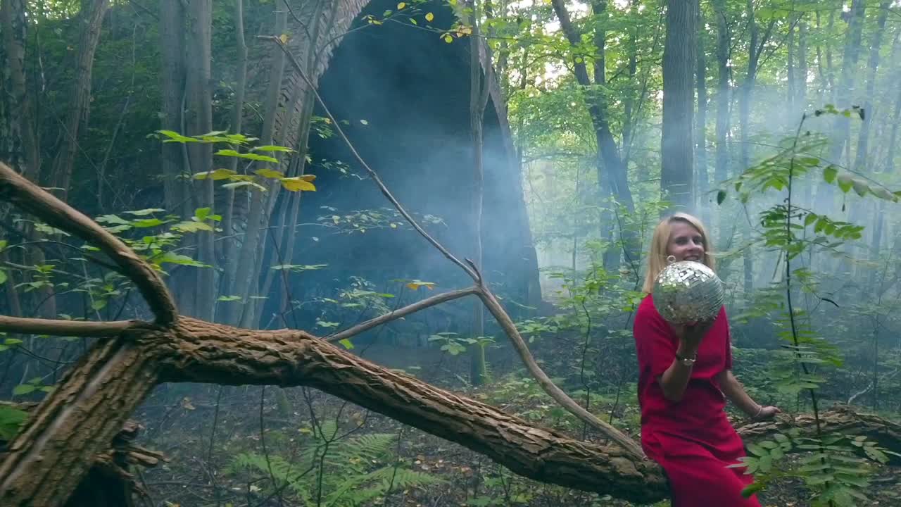 Calmer slow motion cinematic shot in a foggy forest of a young blond girl sitting on a tree with a disco ball in one Hand. Foto video shoot on lost places in wild nature 1080, 24p by Philipp Marnitz