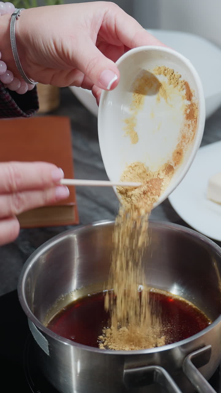 vista parcial de una persona con manga a cuadros vertiendo ingredientes en polvo en una olla de aceite caliente en una estufa eléctrica, con bandeja de madera, especias y utensilios de cocina en la encimera de mármol en la cocina moderna