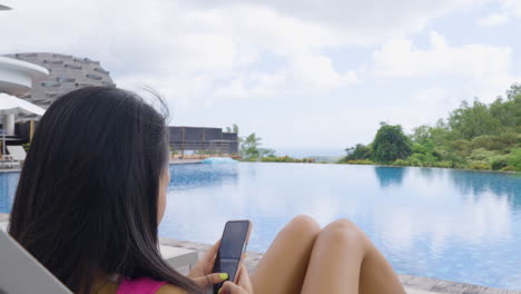 Female-Guest-With-Smartphone-Relaxing-On-Sun-Lounger-By-The-Poolside-In-Hotel