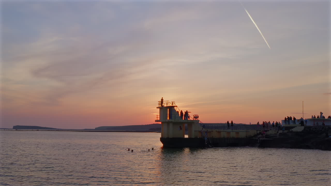 Slow motion aerial static of swimmers at Blackrock Diving Diving Board Salthill promenade coastline in Galway Ireland, gentle waves lapping the shore as dusk settles over the ocean
