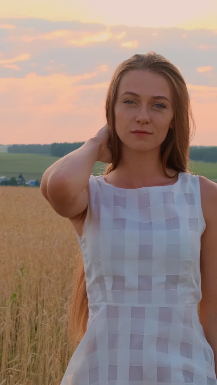 Young woman in a light dress poses gracefully in a golden field during sunset with soft clouds
