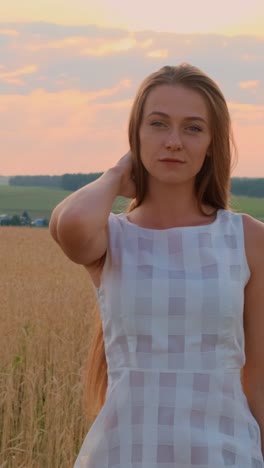 young woman in a light dress poses gracefully in a golden field during sunset with soft clouds