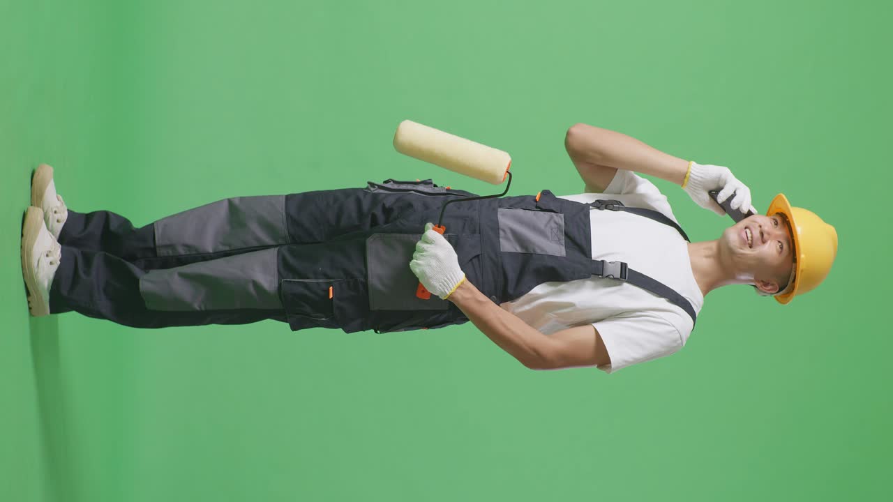 Full Body Of Side View Asian Man Painter Wearing Safety Helmet Talking On Smartphone And Looking Around While Standing In The Green Screen Background Studio