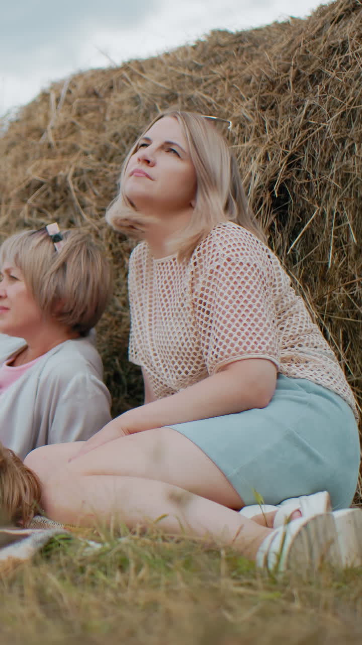 маленькая девочка в розовой рубашке читает свою книгу с зеленой обложкой, лежит на пикниковом одеяле, мать сидит рядом, корректирует волосы, другая женщина отдыхает с собакой на поводке, глядя на расстояние