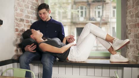 Attractive-young-couple-relaxing-together-at-home-sitting-on-the-window-sill-and-looking-at-each-other.-Beautiful-woman-is-laying-on-the-knee-of-her-boyfriend.-Shot-in-4k