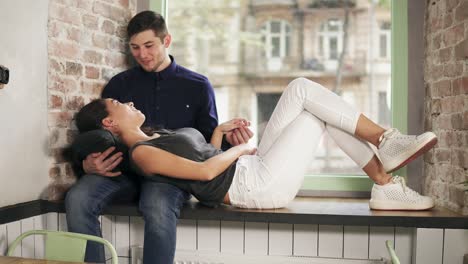 attractive young couple relaxing together at home sitting on the window sill and looking at each other. beautiful woman is laying on the knee of her boyfriend. shot in 4k