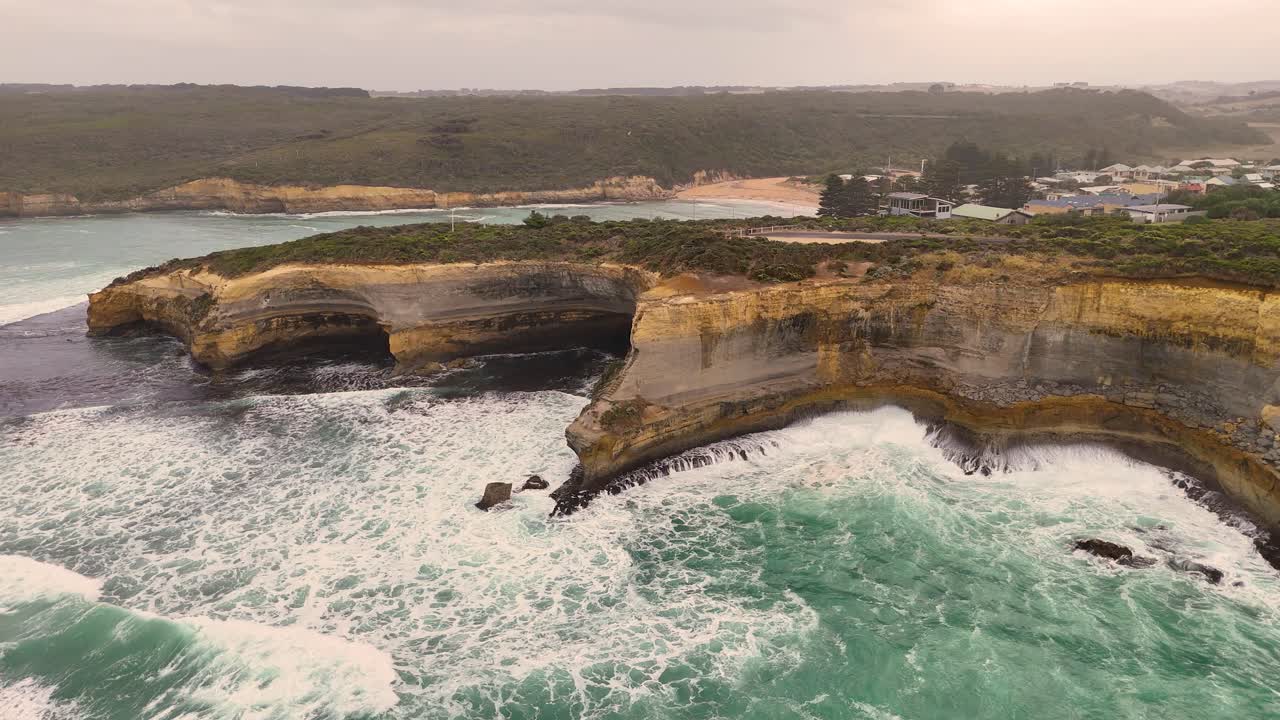 Drone footage captures the dramatic cliffs and turbulent sea at Port Campbell, Australia, under overcast skies with dynamic camera movement