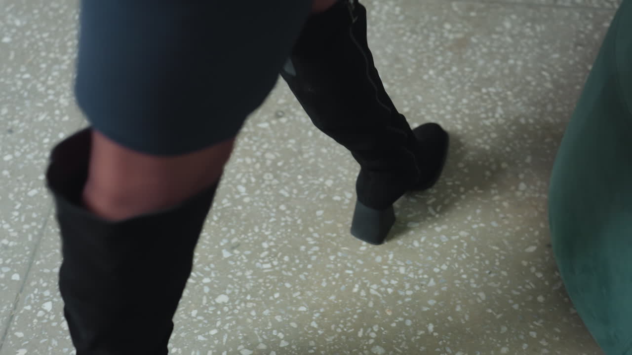 Close up of woman legs in black boots as she walks confidently across terrazzo floor toward green couch in professional office setting, preparing to sit down and begin her workday