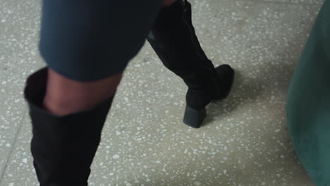 close up of woman legs in black boots as she walks confidently across terrazzo floor toward green couch in professional office setting, preparing to sit down and begin her workday