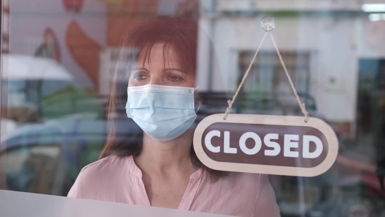 Woman with face mask changing closed to open sign on window looking outside waiting for clients. Start-up at covid time