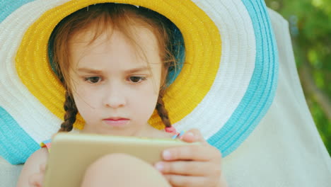 A-Cute-Little-Girl-In-A-Large-Multi-Colored-Hat-In-A-Bathing-Suit-Is-Sitting-On-A-Lounger-And-Playin