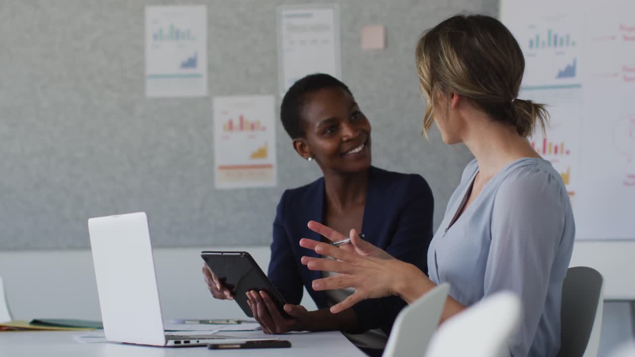 Due colleghe di diversa etnia sedute alla scrivania con un laptop che discutono in ufficio.