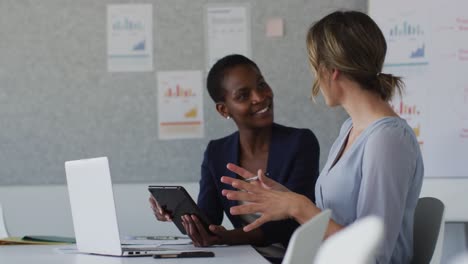 two diverse female colleagues sitting at desk with laptop and discussing in office