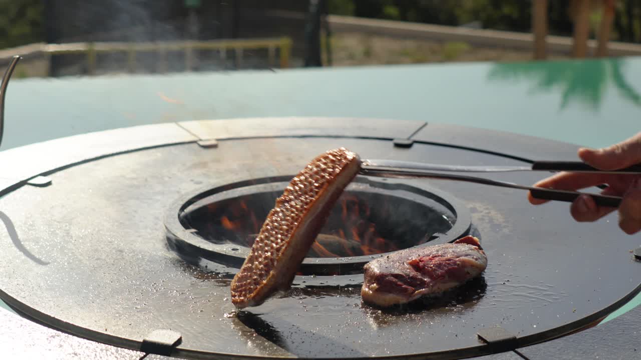 tongs flip a piece of Sizzling meat on a fire pit grill in a scenic outdoor setting