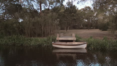 Pequeño-Barco-Amarrado-En-Un-Embarcadero-Rústico-A-Lo-Largo-De-La-Orilla-De-Un-Río-Con-árboles