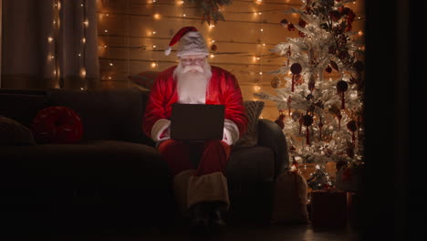 general plan front view a real santa claus is working with a laptop at night with glasses in the light of christmas lights on the background of a christmas tree. remote work