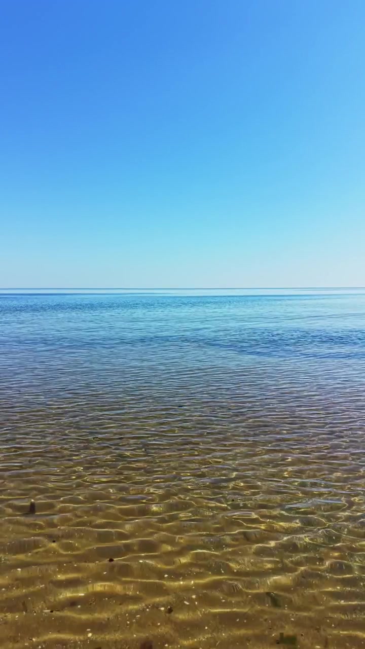 Kirkas sininen taivas ja tyyni vesi ulottuvat rantaa pitkin, kun auringonvalo heijastuu lempeistä aalloista rannalla keskipäivällä.