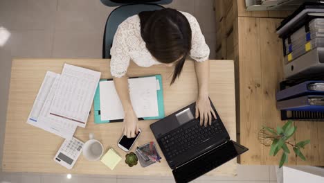 fast forward with top view busy asian businesswoman doing financial paperwork with a laptop is writing note, answering phone and using calculator in the office.