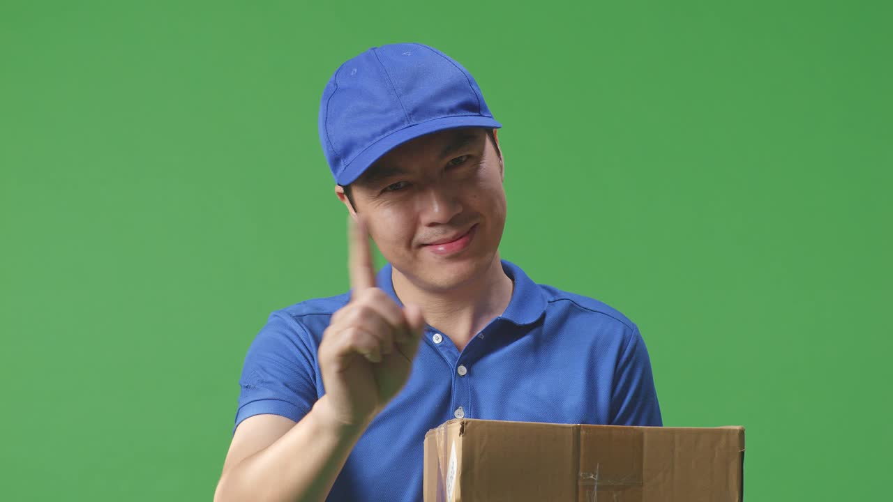 푸른 유니폼을 입은 아시아 남성 택배사의 클로즈업은 미소 짓고 스튜디오에서 녹색 화면 배경에 상자를 배달하는 동안 제스처를 표시하지 않습니다.