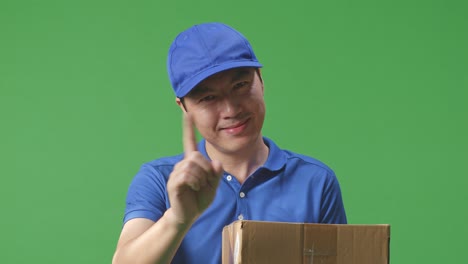 primer plano de un mensajero masculino asiático en uniforme azul sonriendo y sin mostrar ningún gesto mientras entrega una caja de cartón en el fondo de la pantalla verde en el estudio