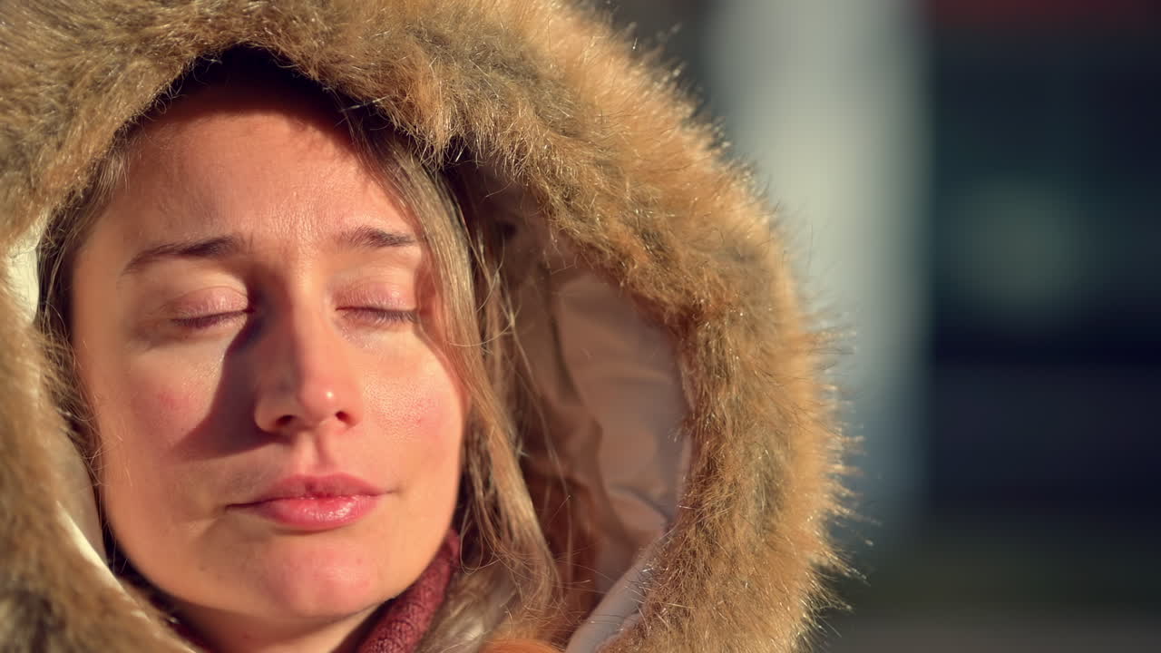 A woman takes a moment to enjoy the warmth of her hooded jacket while outdoors in a cold winter landscape. Her relaxed expression shows comfort against the chill