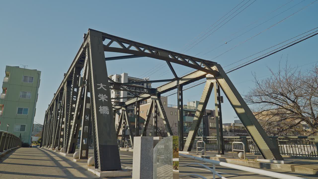 Dark steel truss bridge with Japanese nameplate stands against vivid blue sky in Tokyo, with urban buildings and bare trees alongside, captured as a static low-angle shot from the bridge deck