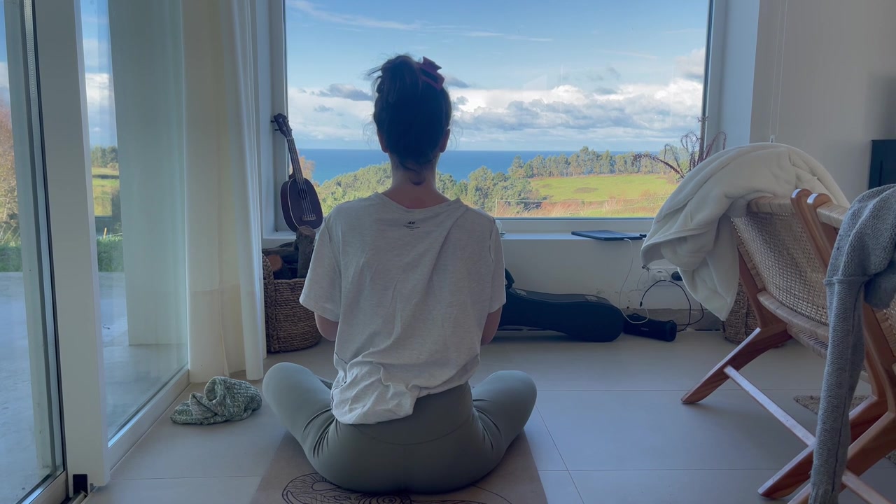 Giovane donna seduta in meditazione su un tappetino da yoga di fronte a una finestra panoramica sul mare