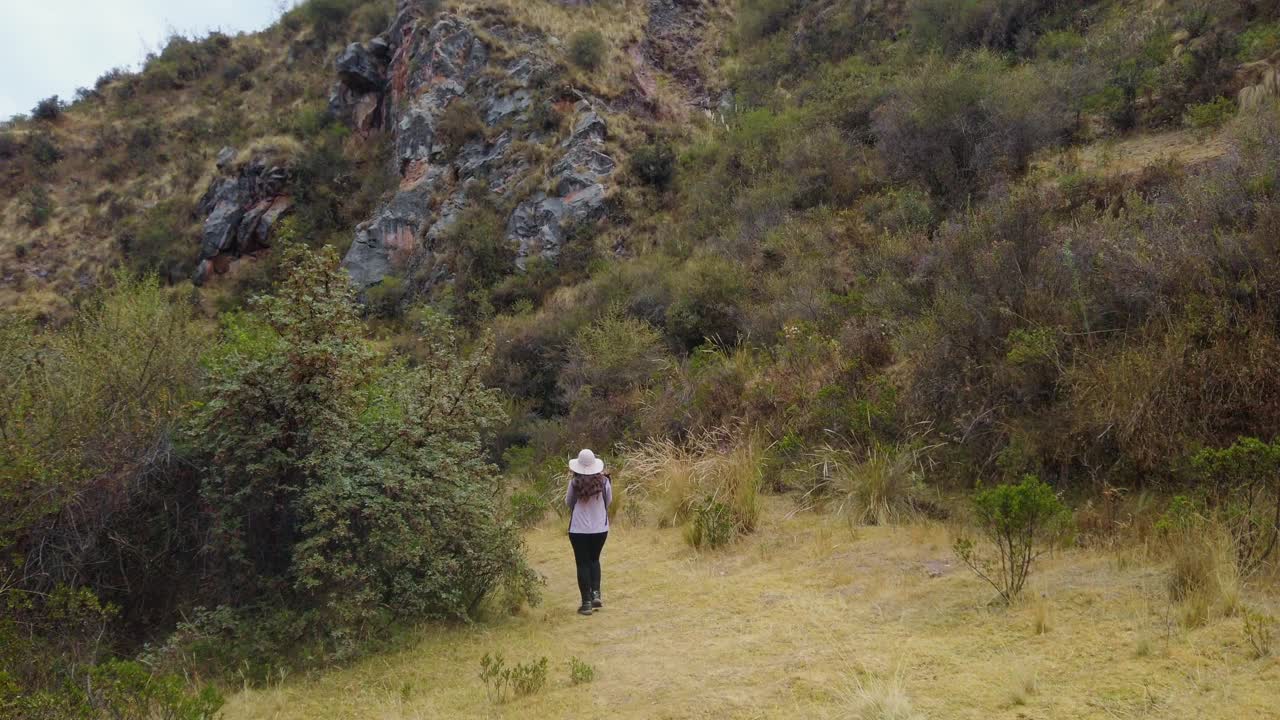 Woman in a hat walks along a lush Inca trail surrounded by mountains near Cusco, Peru leading from Puka Pukara ruins to Inkilltambo