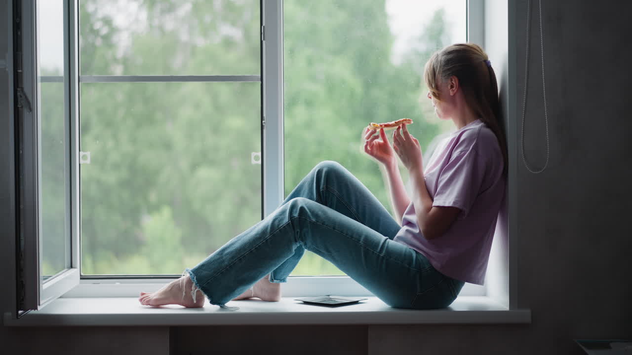 Side view of young woman seated comfortably on window ledge eating pizza slice with plate nearby under soft natural light, peaceful indoor scene with blurred lush green outdoor background of trees