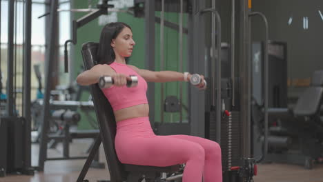 a brunette woman in a pink suit raises dumbbells to the sides while training her shoulders in the gym. seated shoulder and arm bench exercise