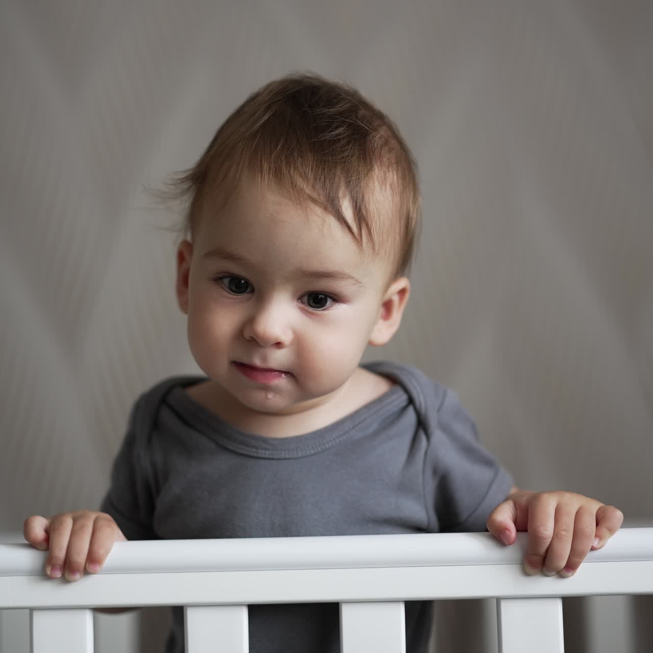 Beautiful kid in grey t-shirt standing in white cot. Smiling baby boy waving his hand sweetly. Close up