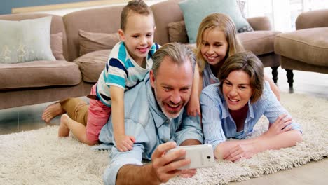 Familia-Feliz-Tumbada-En-La-Alfombra-Y-Hablando-Un-Selfie-Por-Teléfono-Móvil