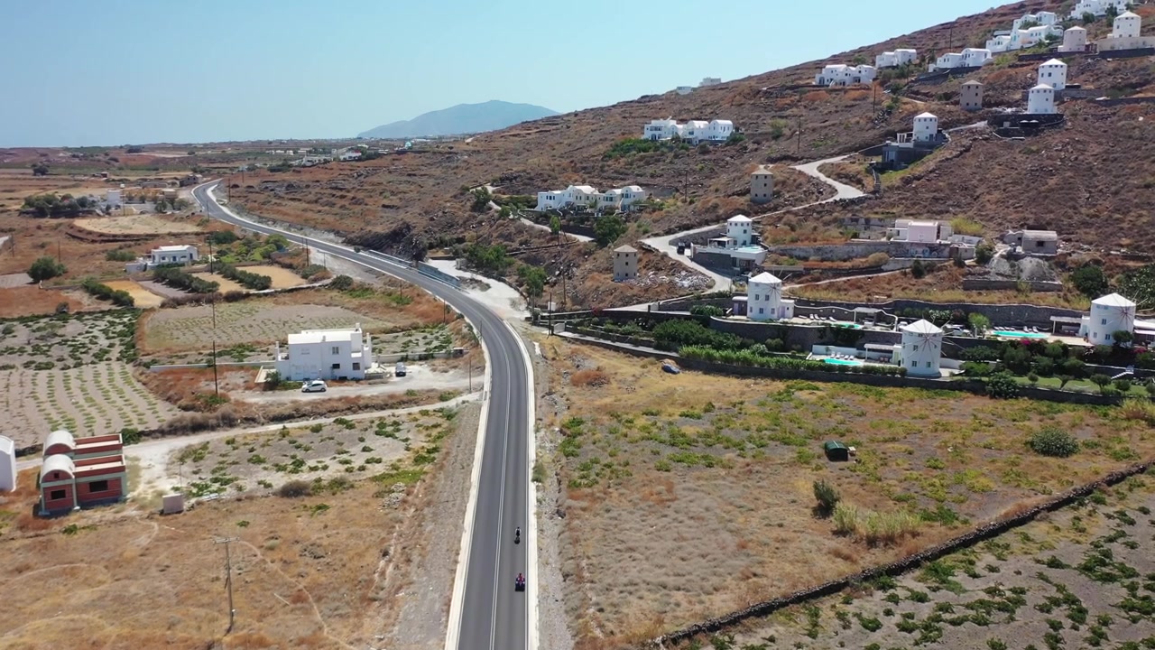 Splendida ripresa con drone che segue due viaggiatori che guidano quad lungo una strada costiera tortuosa a Santorini. Affiancata da aspre scogliere vulcaniche, ville bianche tradizionali e l'oceano blu profondo
