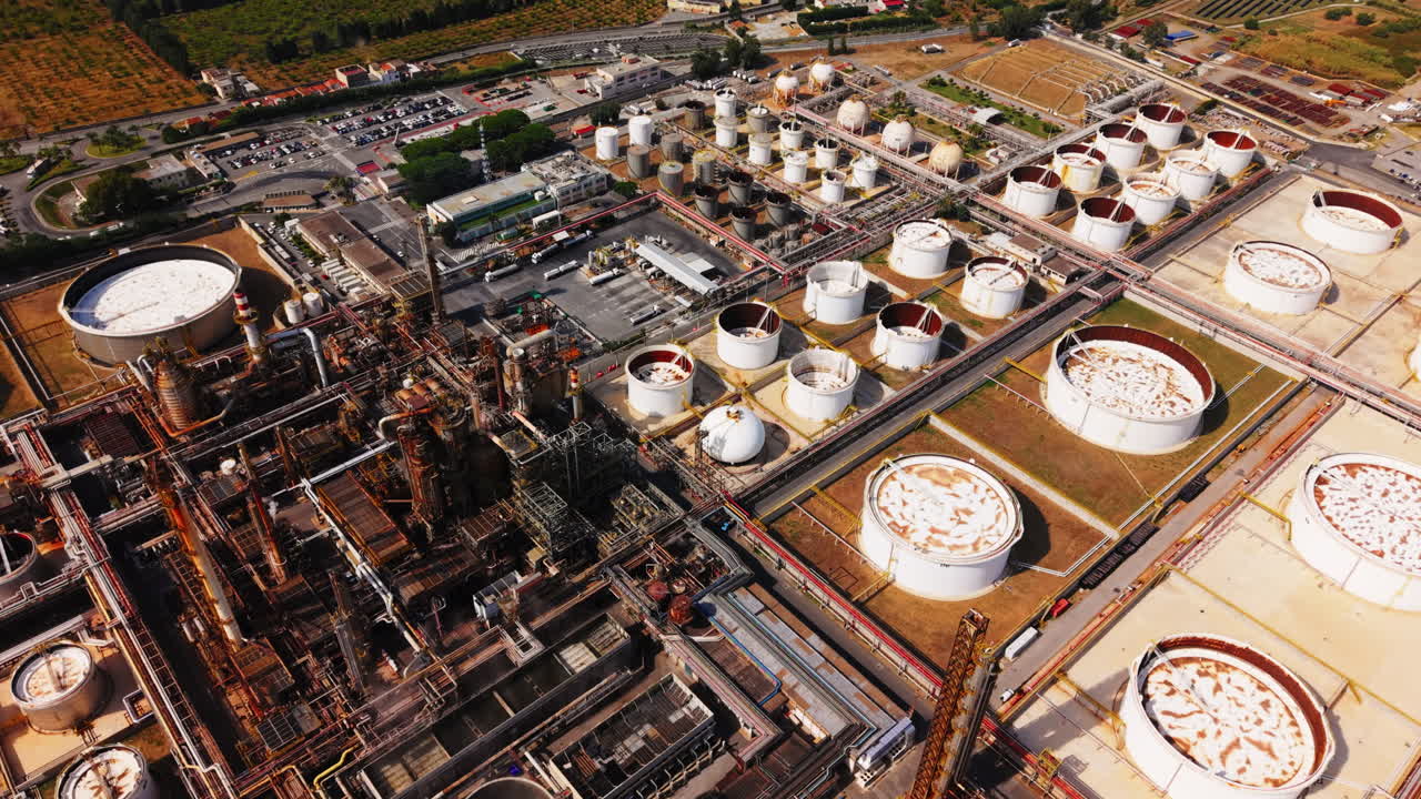 Drone in steep top-down angle capturing oil refinery tanks, pipelines, and surrounding roads in Sicily