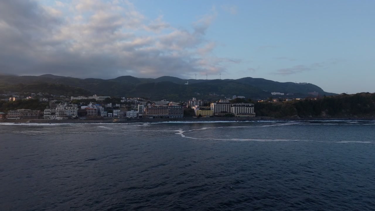 Cittadina costiera fiancheggiata da hotel sotto un crinale boscoso con turbine eoliche a Higashiizu, Izu, Giappone, nella morbida luce del tardo pomeriggio, ripresa aerea con drone
