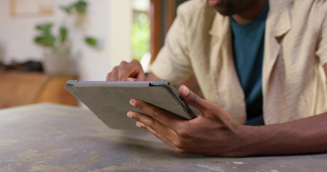 Prohlížení obsahu, dospělý africký muž klepající a posouvající prstem po tabletu v šedém pouzdře u domácího stolu, usmívající se.