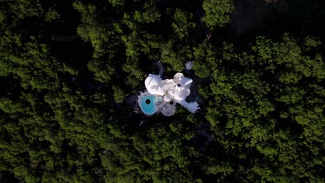 top-down aerial view of a white bubble house in the forest, revealing a circular pool, skylights, chimneys, and round windows as the camera approaches