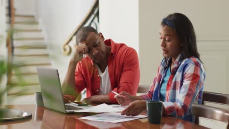 pareja biracial estresada sentada en la mesa con una computadora portátil y contando el presupuesto de la casa