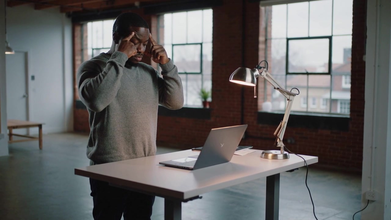Mand i sweater løfter hænderne til tindingerne, sætter brillerne fra sig i et loft, forbereder sig på at arbejde på bærbar computer.