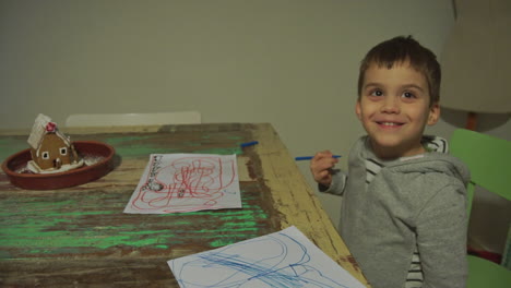 tiro inclinado de un niño pequeño caucásico dibujando con marcadores de color en papel y sonriendo a la cámara