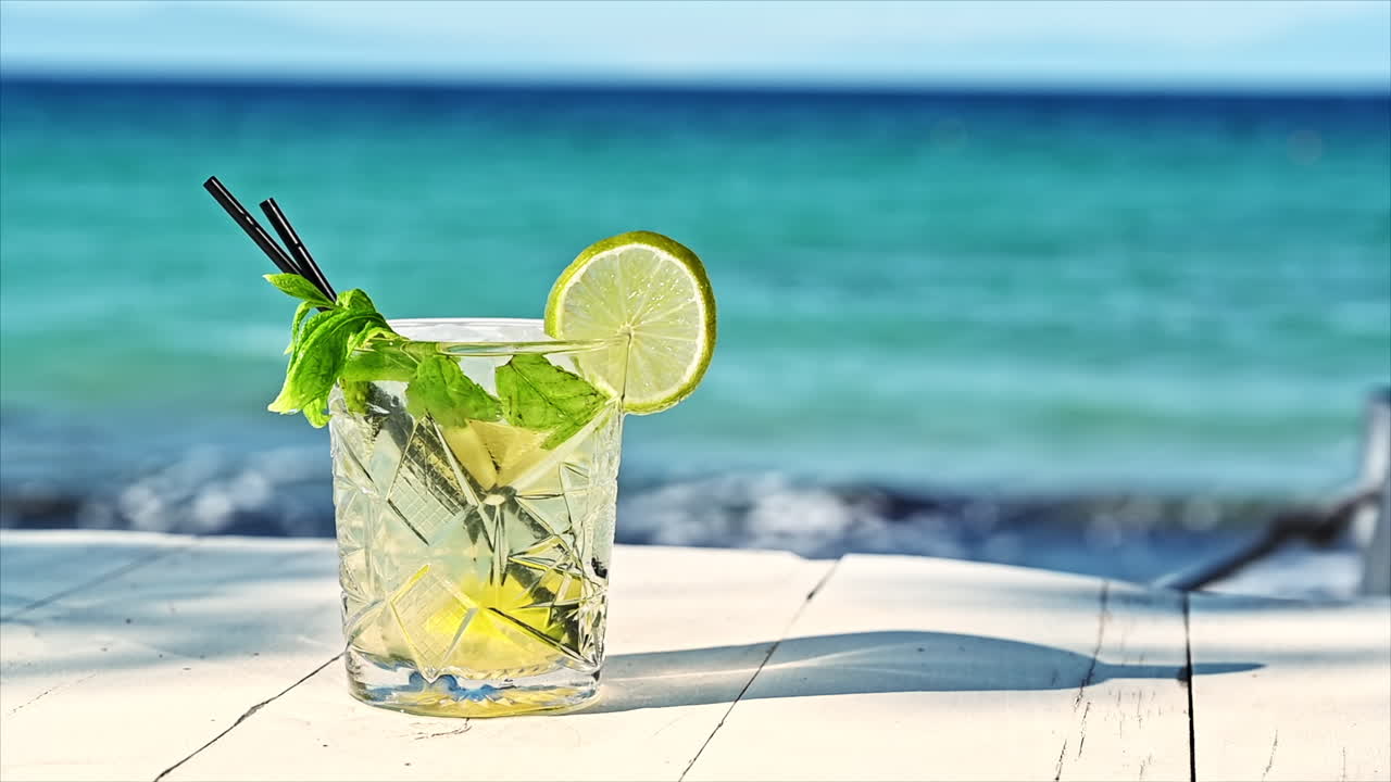 Cocktail with ice, slice of lime, aegean sea on the background, Greece. Slow motion