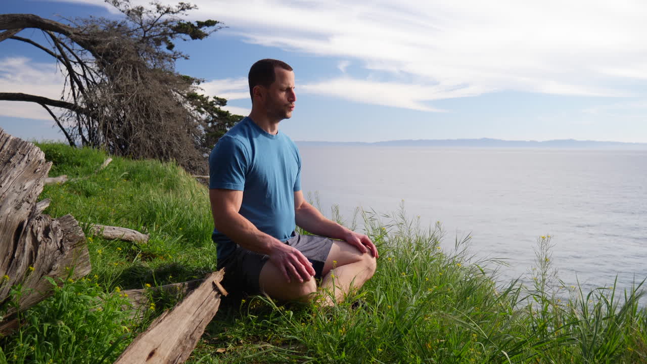 un hombre sentado en una pose de meditación practicando ejercicios de respiración profunda y atención plena en un acantilado de playa sobre el océano azul en santa bárbara, california.