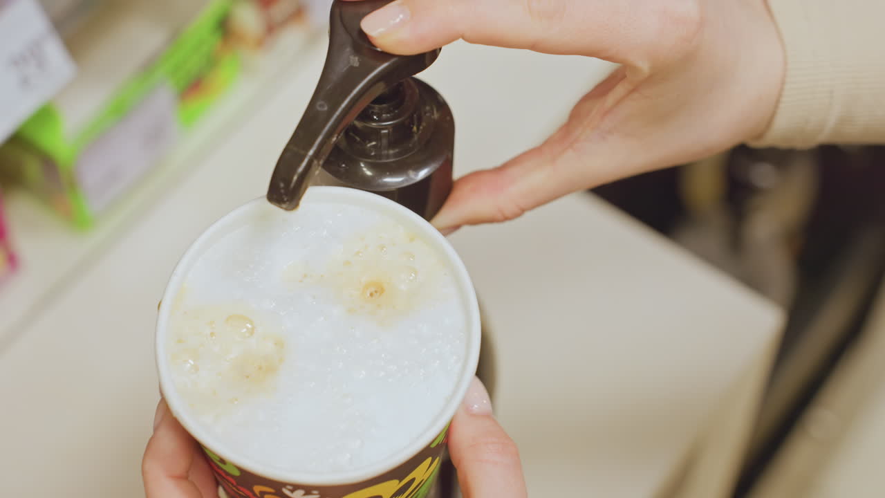 Kafkas barista, köpüğe şurup döküyor. Bir bardaktaki kremalı kahve köpüğünün üzerine karamel gezdirmek için pompa kullanan elin yakın çekimi, sıcak kafe tezgahı, sıcak aydınlatma, dokulu köpük, arka plan ürün rafları