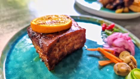 slow parallax effect reveals a tender glazed pork belly topped with orange slice, artful pickled vegetables, and mushrooms on a blue ceramic plate