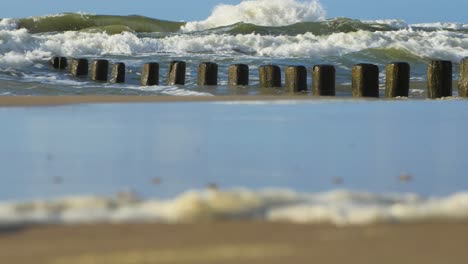 Stormy-waves-breaking-against-old-wooden-pier-on-the-beach,-white-sand-coast,-sunny-day,-Baltic-sea,-distant-low-angle-medium-shot