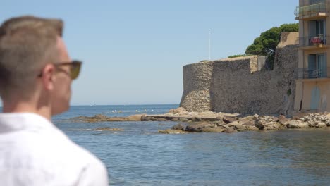 a blurred man from the back walking forward with a view of the sea and a stone construction then a building on the right, the man has short hair and wears his jacket on the shoulder, sunny weather