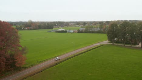 drone shot following silver, grey car between green grass fields on a dutch road, street from above